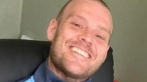 Michael is smiling at the camera and appears to be sat in a chair and wearing a blue top. He has close-cropped hair and a short beard.