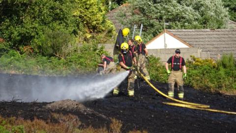 Firefighters direct hoses across a burnt area of heathland with houses in the background