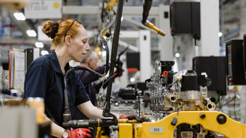 Female JLR worker in a blue polo shirt assembling an engine, with other workers behind her on a production line. Various pieces of equipment are on the line and hanging above