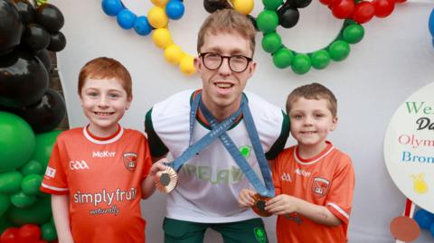 Two-time Olympic medallist swimmer Daniel Wiffen meets Conor, 8, and Matthew, 6, from Stevenson, during his homecoming event in Magheralin. The boys are posing with Daniel's medals which are hanging around his neck. There is a balloon arch in the background.