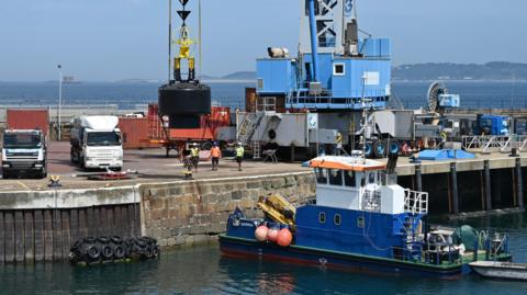 A boat in the water. It is stopped in the harbour next to a brown wall. There is a blue crane holding a black bouy.