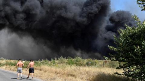 Two men walk past a big grey cloud of smoke, the site of Arnolds Field in Launders Lane, Rainham