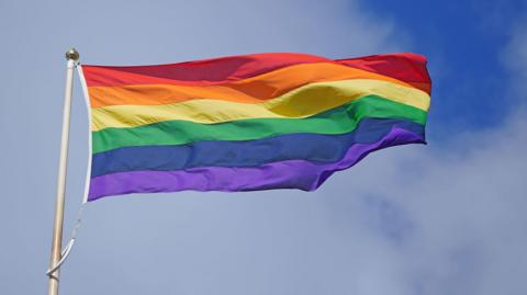 A rainbow-coloured Pride flag flying against a blue sky with light clouds.