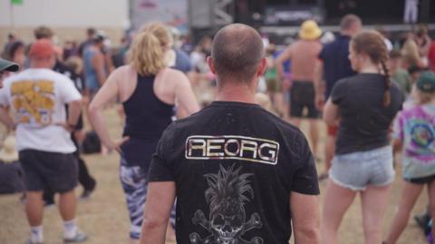 A man is wearing a black t-shirt with the Reorg logo printed on the back. He is facing away. Other people are standing in front of him with their backs to the camera. A stage is in the background.