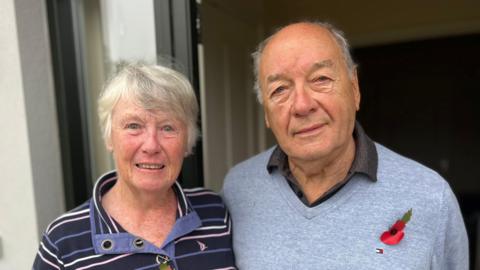 Evie is an older woman with blue eyes and grey hair. She is standing to the left of the photograph, wearing a striped purple and navy blue jumper. To her right is Tony - an older man with a bald head, apart from some grey hair on the sides. He has blue eyes. He is wearing a grey jumper with a grey shirt underneath. He is wearing a poppy. The couple are standing in front of a doorframe.