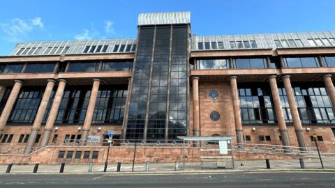 Newcastle Crown Court. It is a large imposing building with red brick walls and columns and large dark windows.