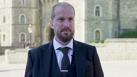 Nathaniel Dye, a man with a beard, in a black suit standing in front of Windsor Castle