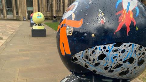 A dark coloured globe depicts images by children of space travel. It is on a plinth outdoors with other globes in the background