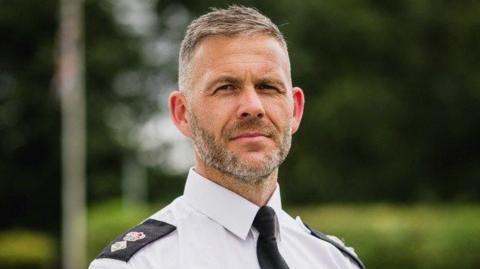 A man who appears to be in his early 50s in a police uniform - white shirt with black shoulder pads and a black tie. He looks at the lens with a neutral expression. He has short greying hair and a short beard.