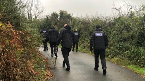 Police officers walking down a narrow country lane. The officers are dressed in black trousers and black jackets with the police logo on the back. They are walking between two hedges.