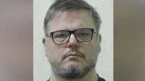 Mugshot of Dean Juric. He has scruffy dark and grey hair and a black and grey beard and is wearing black-framed glasses