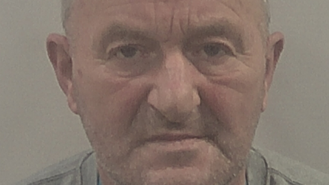 Close-up custody shot of Michael. He has thin receding grey hair and stubbly beard, and is wearing a grey top.