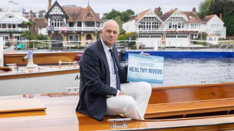 Sir Steve Redgrave in 2023 sits on a polished wooden boat wearing a navy blazer, white trousers, and a tie, holding a sign that reads "I SUPPORT HEALTHY RIVERS." Behind him, traditional boathouses and riverside buildings line the opposite bank of the river under an overcast sky.