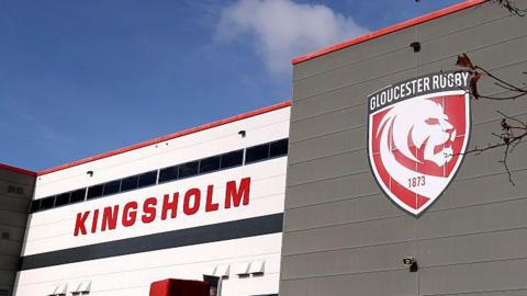 A general view of the exterior of Gloucester Rugby's Kingsholm stadium with the word "Kingsholm" in large red letters on a white background and the club's badge showing a white lion on a red background
