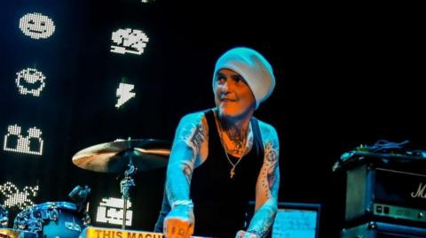 Derry Brownson, the keyboardist of EMF, smiles as he performs on stage. He is wearing a grey beanie, a black tank top and has arms covered in tattoos.