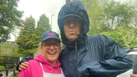 Liam Gallagher in a waterproof coat with arm round woman in lilac top