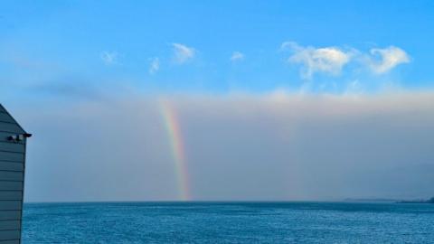 The arc of a bright rainbow in a layer of grey cloud between blue sky and blue sea