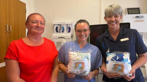 Three women stand in a line in a hospital delivery suite. The woman on the left has a red T-shirt on and her auburn hair is tied back. The other two women are nurses and are wearing blue uniforms. They are holding packages containing clothes and nappies for new babies.