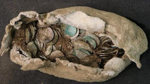 The cloth package containing hundreds of historic coins rests on black surface. The coin faces are detailed while some earth and dirt rest inside the package.