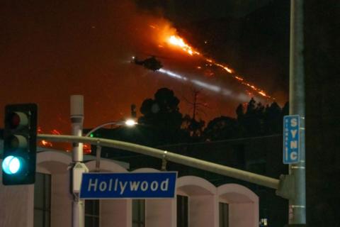 Hollywod street sign, behind it is a helicopter and raging fire.