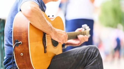 Adult male musician playing a folk acoustic guitar with pickup and cable connected. Performing music outdoors for entertainment.