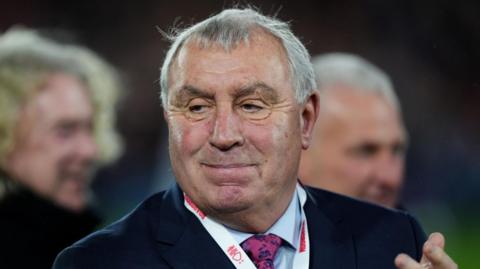 Peter Shilton has short grey hair and is wearing a suit. He is clapping.