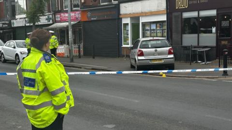 A silver car has mounted the pavement outside a cafe. There is a blue and white police cordon around the scene. There is an police officer wearing a yellow hi-visibility jacket and black trousers, with her brown hair tied in a bun, standing with her back to the camera to the left of the scene. There are two police officers in the same uniform, also wearing hats, standing talking in the distance. 