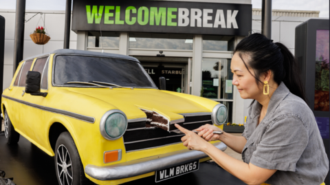 A life-like car made of cake outside a Welcome Break service station. The car's design is based on an Austin/Morris from 1960 and is yellow in colour. A woman with black hair and wearing a grey top is cutting a slice out of the bonnet.