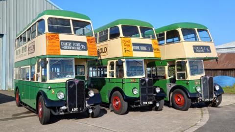 The three classic double decker buses are lined up outside a barn. The sky is blue and the buses are painted green and white, while the wheels are bright red.