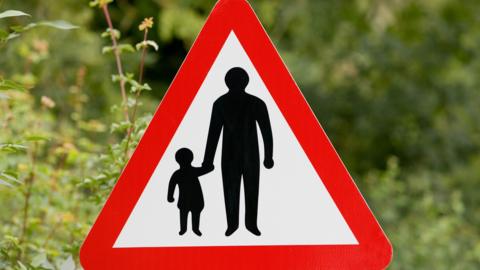 A red and white triangular road sign with a grown-up holding a child's hand 