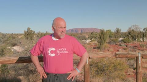 A bald man wearing a pink Cancer Research UK shirt and grey jogging bottoms stands in front of Uluru (Ayers Rock) in Australia. The large monolith is visible in the distance with several trees and patches of sand in front of it. The man has his hands on his hips and stands in front of a wooden fence.