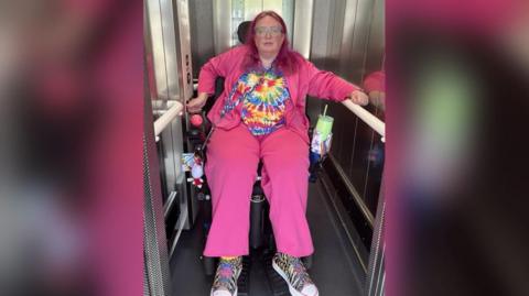 A woman in a bright pink outfit is in a lift in a wheelchair. 