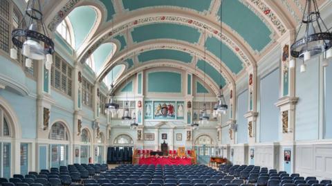 A central hall with pale blue and green walls and ceiling with blue chairs and ornate plasterwork 