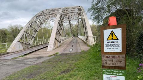 A bridge with a span over it is divided into two paths for cycling and walkers. There is a warning sign asking people not to jump off the bridge 