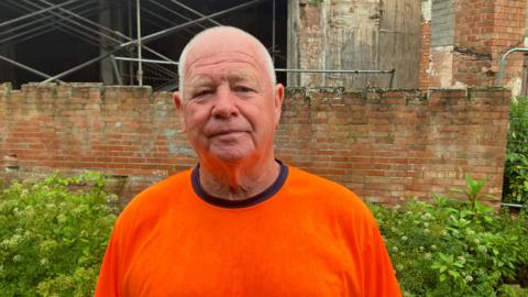 Keith Jackson wearing a bright orange T-shirt looking at the camera with a neutral expression. He is in front of The Life Factory, which is a red brick building in a dilapidated state. There is a hole in the side of the building and scaffolding can be seen through it.