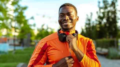 A man zips up his orange sports jacket, he's smiling and wearing headphones around his neck.
