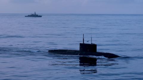 A submarine is seen in the water in the foreground, with a navy warship in the background