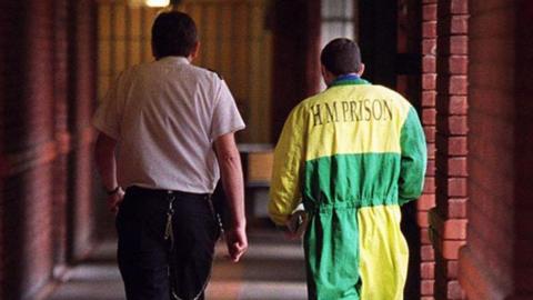 A prison officer in a white shirt and black trousers walking down a corridor alongside a prisoner in a yellow and green jumpsuit with H M Prison printed on the back