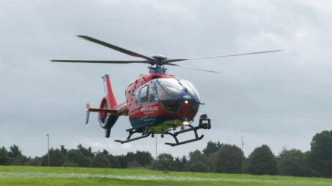 A red and blue Devon Air Ambulance helicopter comes in to land on a grassy field. It has four blades. There are trees in the background. The sky is grey.