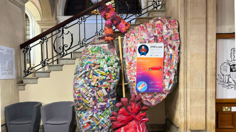 A sculpture of a pair of lungs. The two lungs are made up of hundreds of empty boxes from seized vapes. The left lung has lots of different colours and the right lung is mainly pink. There is also a sculpture of a heart underneath the lungs. 