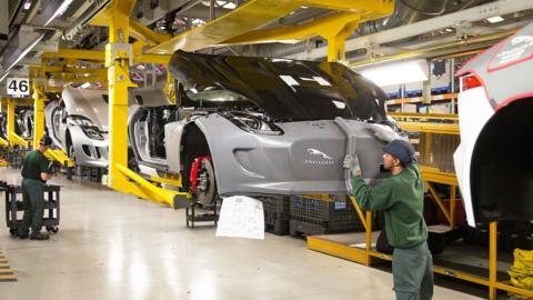 A man in a green jumper and blue cap closely inspects the grey body of a car which is suspended above a factory floor.