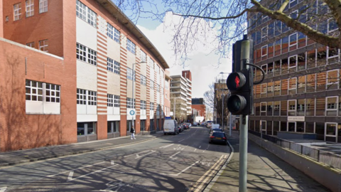 A Google view of Wollaton Street, in Nottingham. On the left a modern three-storey redbrick building can be seen, with an older six-storey building to the right. A traffic light is in the foreground and a number of parked vehicles can be seen. 