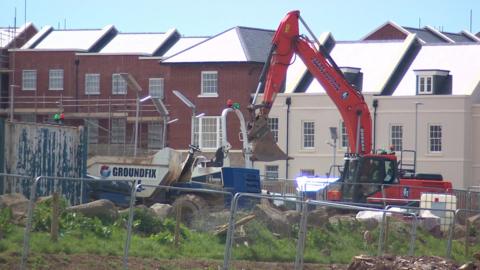 A red digger and a white truck working on a fenced construction site. There are multiple new looking residential homes with some scaffolding in place.
