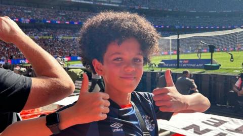 Noah is smiling at the camera during a football match and giving a big "thumbs up" with both hands. He is wearing a dark blue football shirt and the pitch and other fans are in the distance
