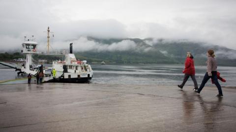 Two women walk down a slipway towards the MV Corran, a white and black ferry with some cars on its deck. It is a rainy day with low cloud on hills on the opposite shore of a loch.