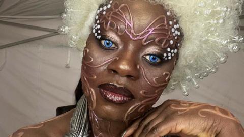 A person in bold avant‑garde makeup with bright blue eyes, a large white beaded wig, and metallic costume accessories, standing in a tent-like backstage setting