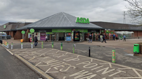 A wide view shows the entrance of an ASDA supermarket, with the green ASDA sign above the main doors and several shoppers walking near the entrance. The foreground includes a marked pick-up point area with painted road text and green bollards lining the pavement. The store is a low brick building beneath a cloudy sky, with parking areas and a few trees visible around the site.