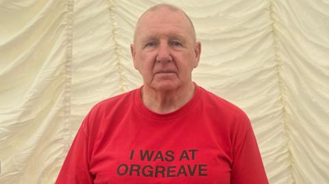 Robert Young, a man in his 80s with shaved hair, is wearing a bright red shirt which reads in black lettering: "I was at Orgreave."