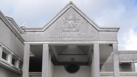 The front of Truro Crown Court. It is grey stone building with COURTS OF JUSTICE written below an elaborate heraldic shield. There are four pillars and the roof is pitched to a point.