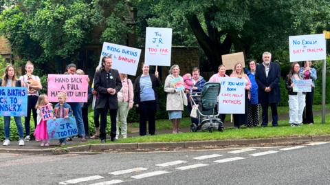 Protesters, including men, women, and children, with placards by the side of the road. One says: 'Bring births back to Banbury.' 